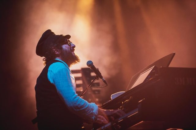 Vinicio Capossela sul palco che suona il Pianoforte durante un concerto, circondato da luci calde